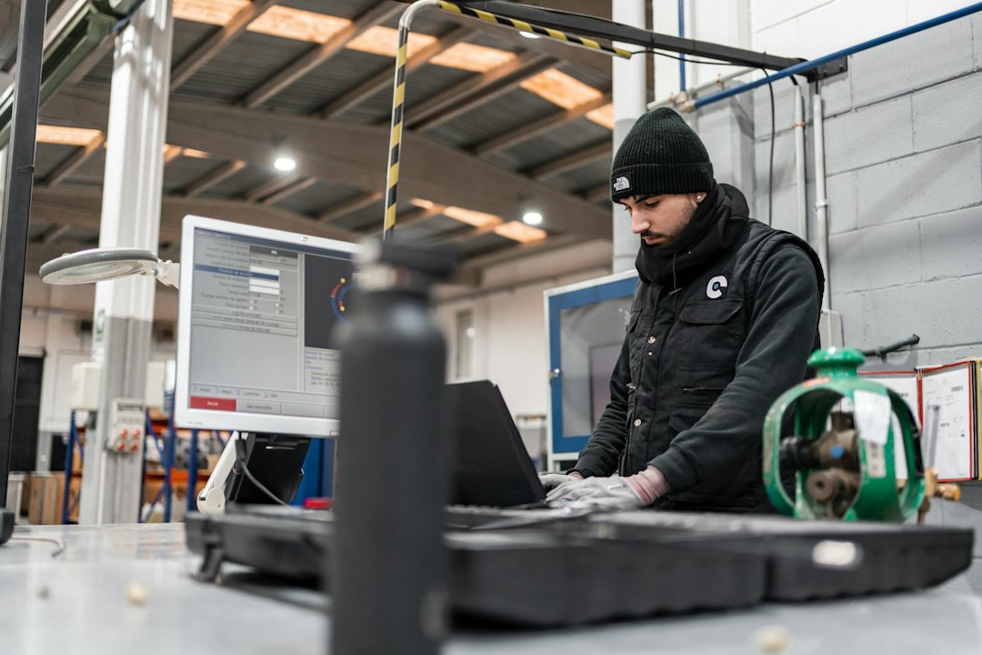 An engineer in a beanie works at a computer in a manufacturing facility with a laptop and industrial equipment