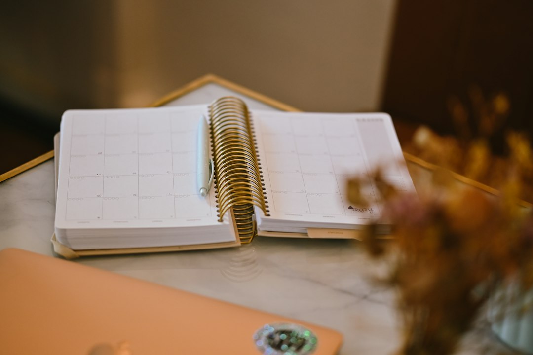 An open spiral notebook with a pen lies on a marble surface beside a small vase of dried flowers