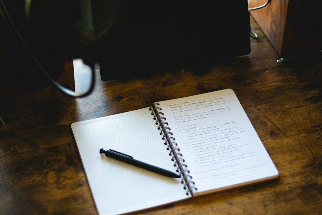 An open spiral notebook with a pen rests on a dark wooden desk in front of a computer monitor